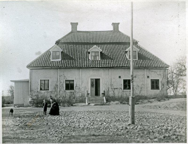 En herrgårdsbyggnad. En kvinna, ett barn och en hund på gården.