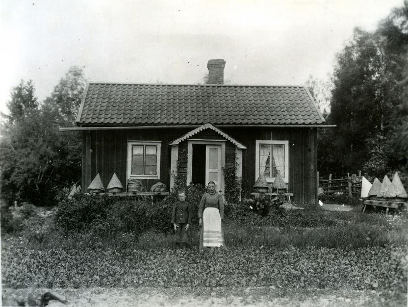 En kvinna och en pojke utanför en trästuga med bikupor framför.