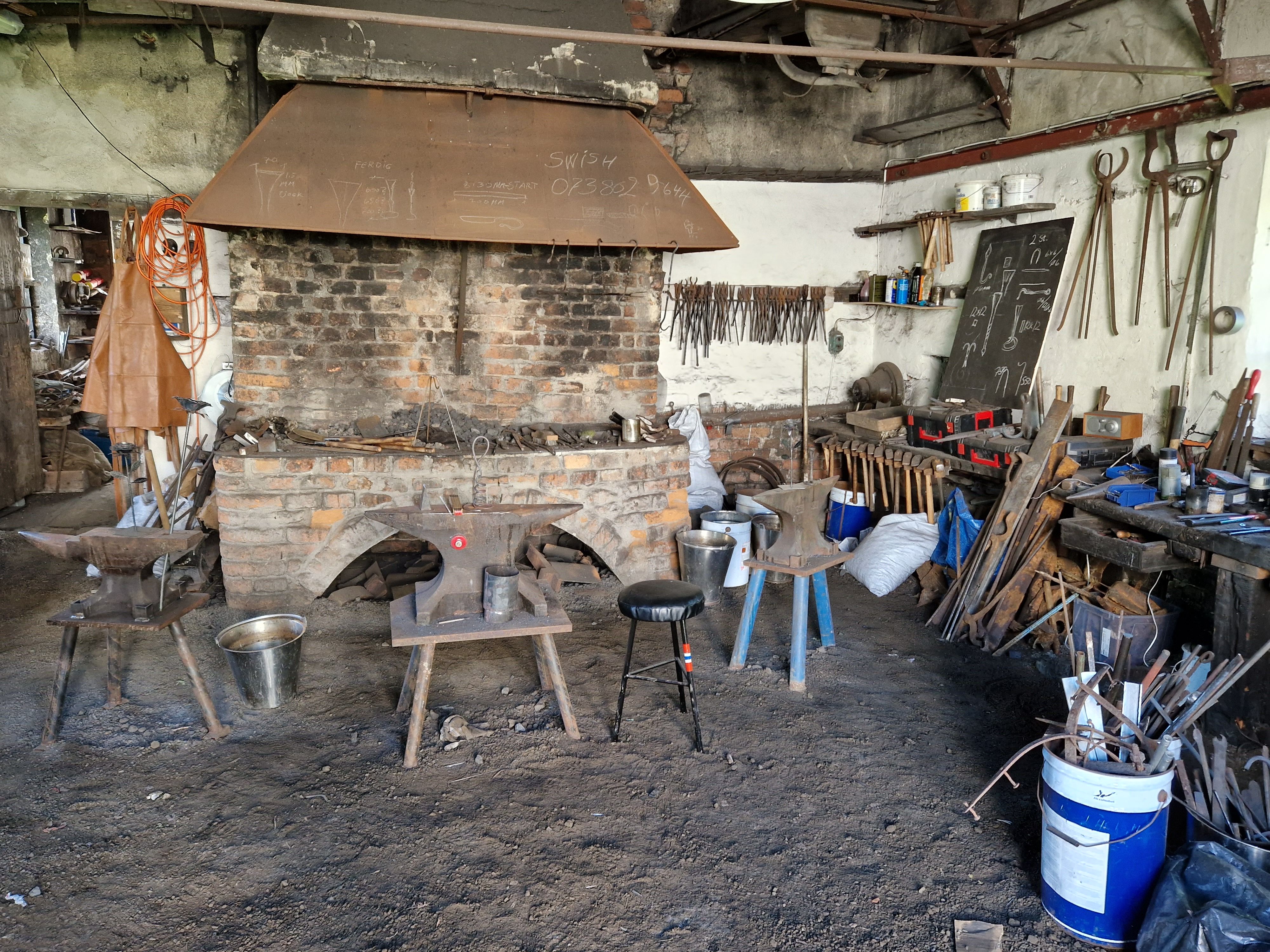 Bilden visar interiören i en smedja för mindre föremål. En härd, ett städ och massor med tänger.