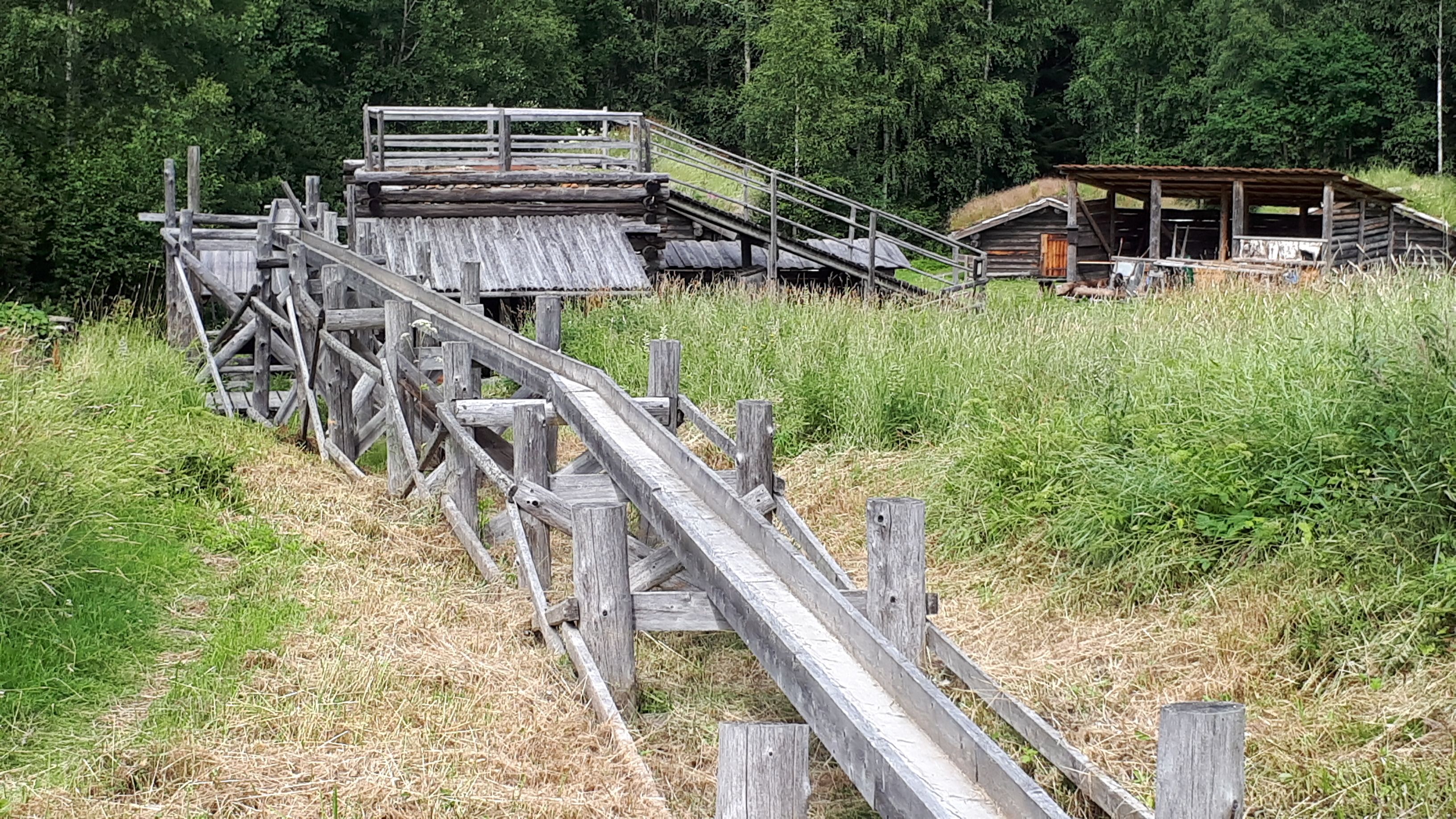 Bilden visar en vattenränna i trä som leder till ett vattenhjul vid den låga masugnen, uppbyggd av trä. Mycket gräs runtom.