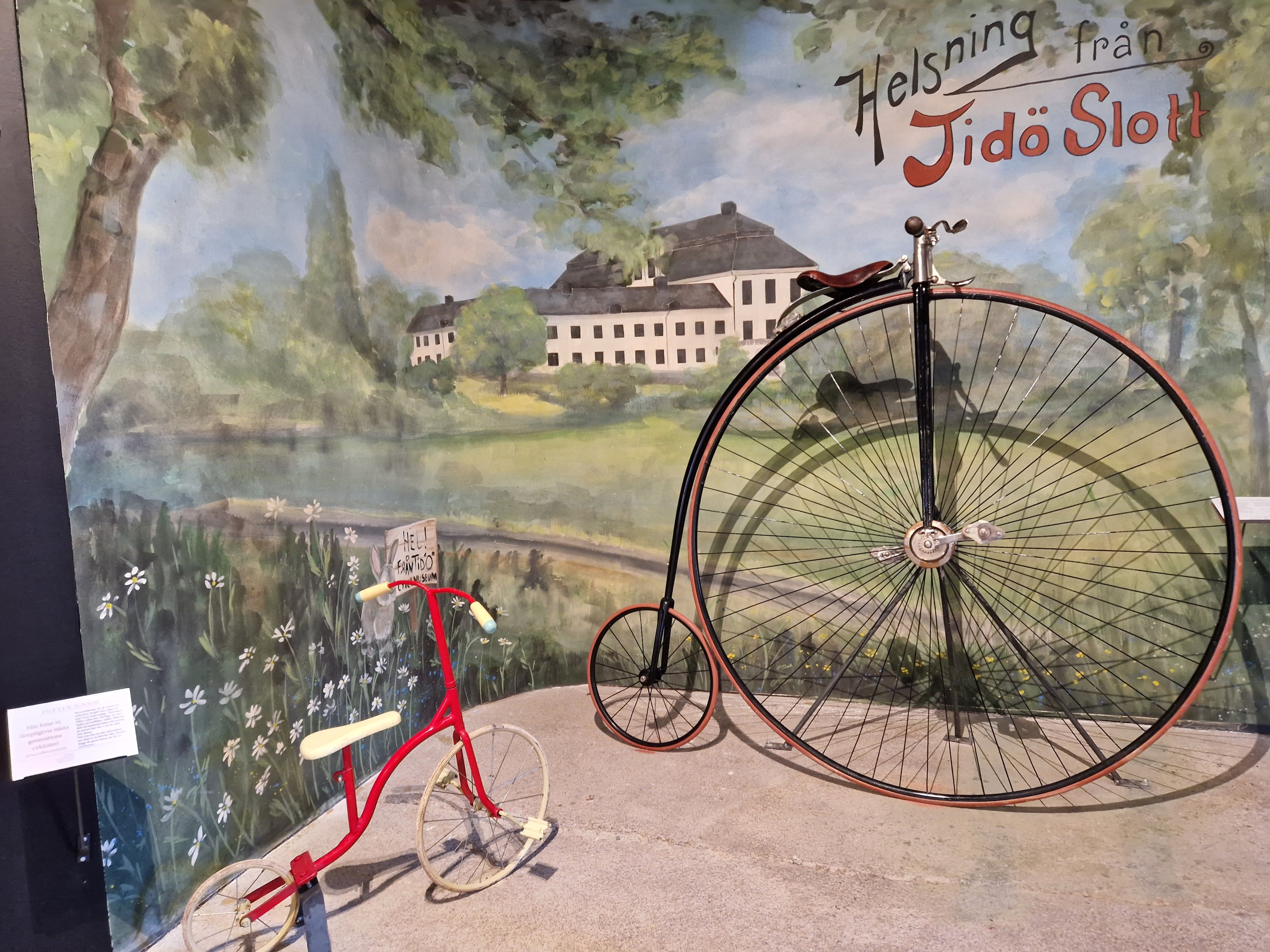 Bilden visar en gammal höghjuling, en cykel, och en barncykel. I bakgrunden en stor bild på Tidö slott.