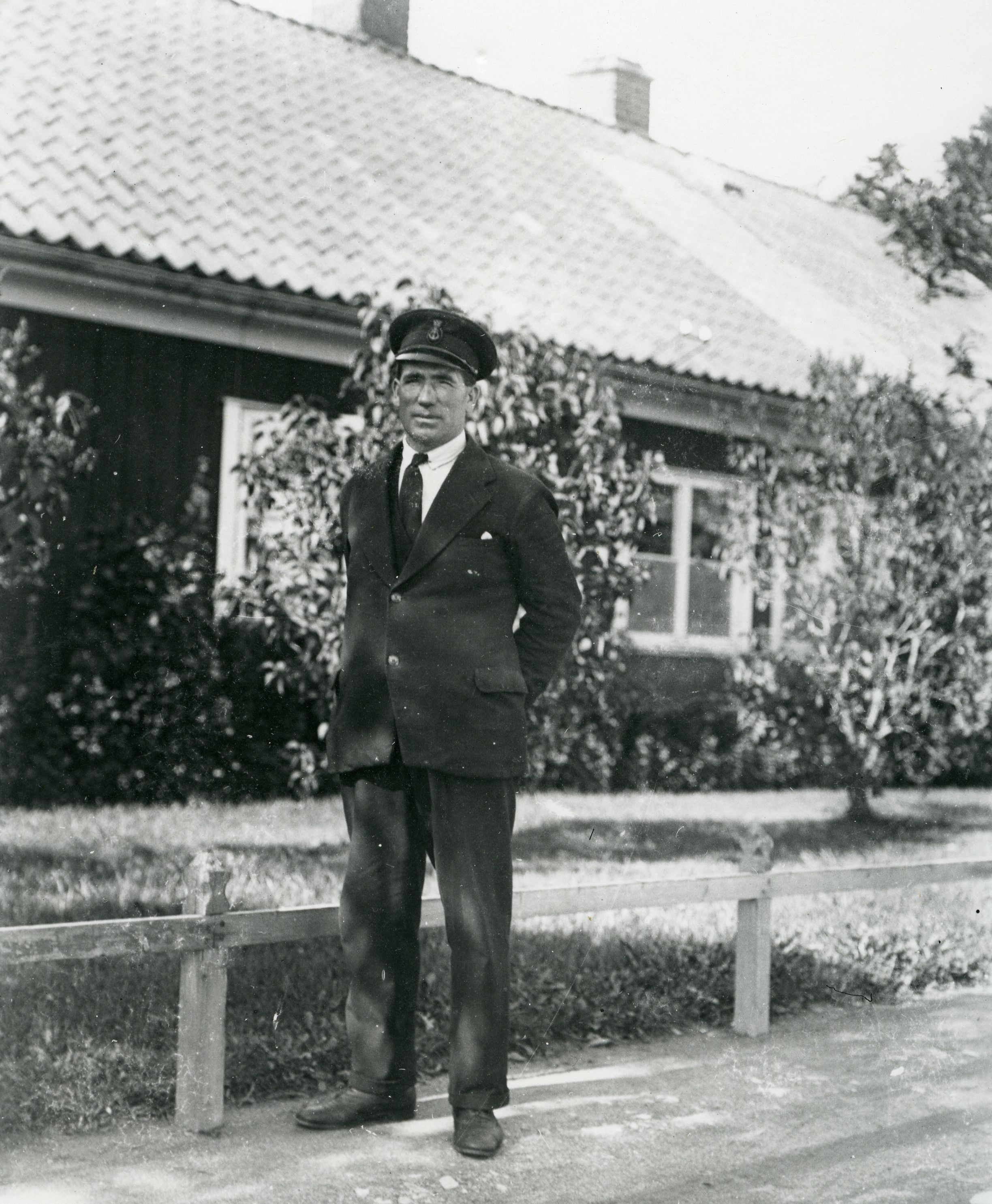 En man i mörk kostym och skärmmössa står framför ett trähus med trädgård, avskärmat av ett staket.